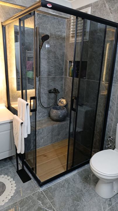Gri mermer görünümlü fayanslı banyo, siyah çerçeveli duşakabin ve taş kurna