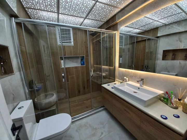 Ahşap ve gri mermer görünümlü fayanslı banyo, arkadan aydınlatmalı ayna ve sürgülü cam duşakabin